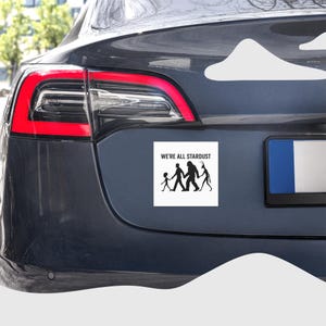 May include: A white rectangular car magnet with the text "WE'RE ALL STARDUST" above a black silhouette of a family and a Bigfoot-like figure. The magnet is affixed to the rear of a dark gray car.