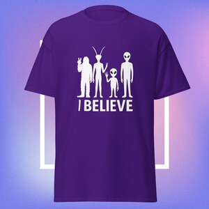 May include: A purple t-shirt featuring a white graphic of four alien figures and the text "I BELIEVE". The figures include a Bigfoot-like creature, two tall aliens, and a small alien. The shirt is a solid color.
