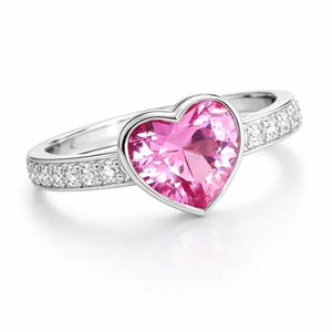 May include: A silver ring featuring a large, heart-shaped pink gemstone. The band is adorned with small, clear gemstones. The ring is set against a white background, highlighting its design and sparkle.