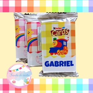 Può includere: Tre Kinder Cards confezionate singolarmente. Le confezioni presentano un trenino dei cartoni animati, la scritta "Kinder Cards" e il nome "Gabriel". Altre confezioni hanno un design arcobaleno e stella. Sfondo colorato.
