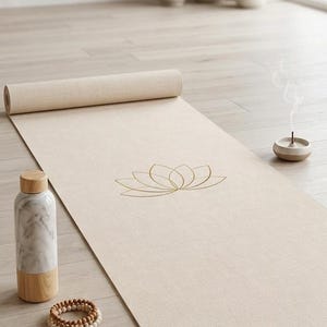 May include: Beige yoga mat with a gold lotus flower design, rolled up at one end. A marble water bottle, wooden beads, glasses, and a small incense burner are nearby. The scene is set on a light wood floor.
