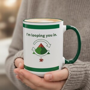 以下が含まれることがあります： 緑色のハンドルと縁が付いた白いセラミックマグ。テキスト「I'm looping you in.」と矢印付きの山のグラフィックが特徴です。小さなコーヒーカップのグラフィックが下にあります。