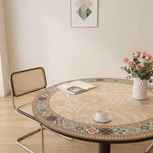 May include: A round dining table with a floral patterned top, a decorative border, and a black base. A cup of coffee, a book, and a vase of pink roses are on the table. A chair with a woven back is next to the table.