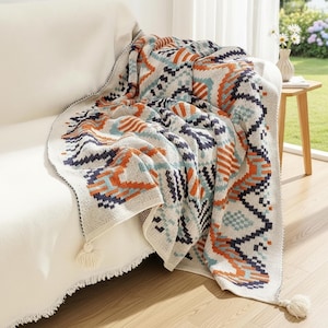 May include: A cream-colored knitted throw blanket with a geometric pattern in shades of orange, teal, and navy blue. The blanket is draped over a white sofa, with tassels on the corners. The blanket's dimensions are not visible.
