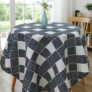 May include: A round tablecloth with a geometric pattern in navy blue, white, and gray covers a wooden table. The pattern features squares and stripes. A light blue vase with greenery sits in the center. The table is in front of a window.