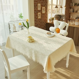 Puede incluir: Un mantel rectangular color crema con un patrón texturizado y bordes con flecos cubre una mesa blanca. Una taza de té amarilla y un plato pequeño con un pastel están sobre la mesa. También se ve un jarrón con tulipanes amarillos y una silla blanca.