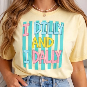 May include: A light yellow t-shirt with the text "I DILLY AND I DALLY" in pink, blue, and yellow. The text is set against a background of vertical teal stripes.