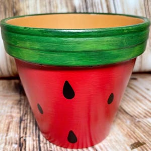 May include: A hand-painted ceramic flower pot with a watermelon design. The pot is red with black seed shapes and a green rim. The interior is a light orange color. The pot is designed for planting flowers or herbs.