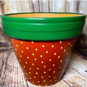 Könnte beinhalten: Handbemalter Blumentopf aus Terrakotta. Der Topf ist rot mit gelben Punkten, einem grünen Rand und einem orangefarbenen Innenraum bemalt. Er ist für Pflanzen und Blumen gedacht.