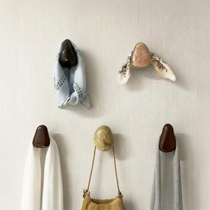 Pode incluir: Uma coleção de ganchos de parede decorativos em várias formas e acabamentos. Alguns ganchos seguram lenços e uma bolsa. Os ganchos têm um acabamento castanho escuro brilhante e um aspeto de pedra natural.