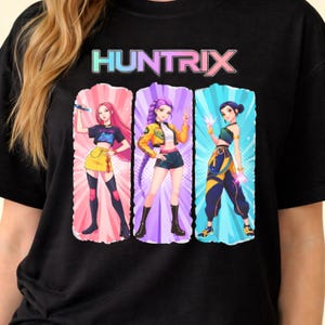 Puede incluir: Camiseta negra con la palabra "HUNTRIX" en una fuente colorida sobre tres figuras de estilo anime. Cada figura está en una pose diferente y lleva un atuendo único, sobre un fondo rosa, morado y azul.
