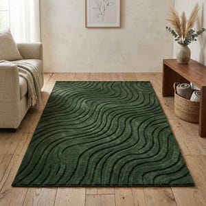Op de afbeelding: Een donkergroen vloerkleed met een golvend, getextureerd patroon. Het rechthoekige kleed bedekt een groot deel van de houten vloer. Het ontwerp creëert een gevoel van diepte en voegt een moderne touch toe aan de kamerinrichting.