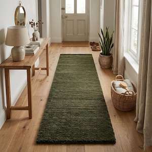 May include: A long, olive green runner rug in a hallway. The rug is made of a textured material and is placed on a wooden floor. The hallway also features a wooden console table, a lamp, and a woven basket.