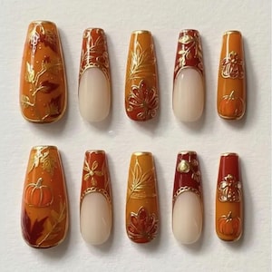May include: A collection of ten artificial nails with autumnal designs. The nails are in shades of orange, red, and gold, decorated with leaf and pumpkin patterns. Some nails have a nude base with gold edging.