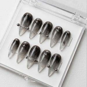 Può includere: Un set di dieci unghie finte in una custodia di plastica trasparente. Le unghie presentano un design sfumato, che passa da un grigio scuro a una punta trasparente. Ogni unghia è decorata con elementi metallici color argento, tra cui barbell e borchie.