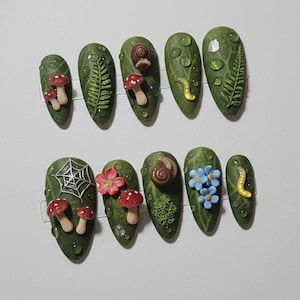 May include: A set of ten green, almond-shaped artificial nails with a forest theme. Each nail features 3D embellishments including mushrooms, snails, ferns, and water droplets. Some nails have a spiderweb or small blue flowers.