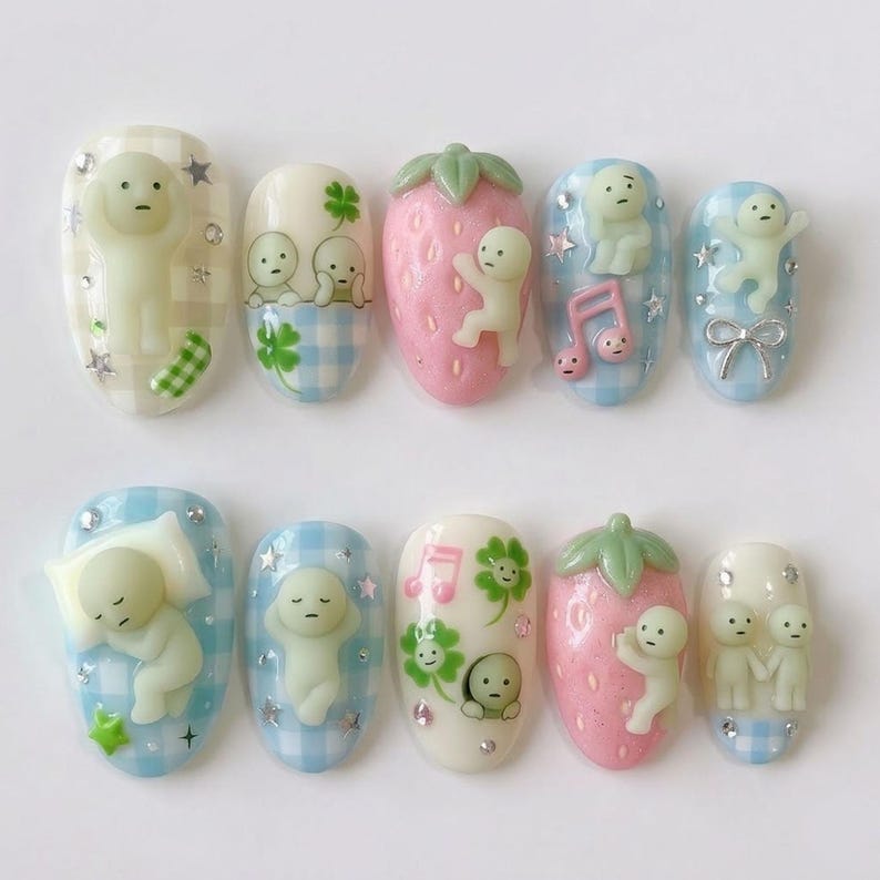 May include: A set of ten artificial nails with various designs. The nails feature a light green, cartoon-like figure, strawberries, musical notes, and four-leaf clovers. The base colors include white, pink, and blue gingham patterns, with silver star accents.