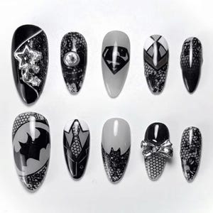 May include: A set of ten artificial nails with various designs. The nails feature black, gray, and white colors with superhero logos, lace patterns, and silver embellishments. The nail shapes are oval.