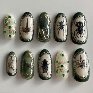 May include: A set of ten artificial nails with unique designs. The nails feature insect and nature-inspired art, including dragonflies, spiders, and centipedes, set against a cream or green background. Some nails have a metallic gold or green finish, while others have green gem accents.
