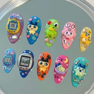 May include: Ten brightly coloured sculpted nail art designs. Each nail features a different 3D character or design, including a yellow Tamagotchi, a blue duck, and a sleeping creature. The nails are set against a reflective surface.