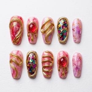 May include: Ten artificial nails with a marble effect base in pink and red hues. Each nail is adorned with gold embellishments, including swirls, gems, and glitter, creating a luxurious manicure design. The nails are presented on a white background.