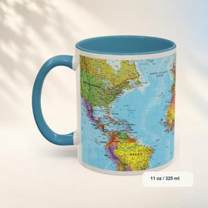 May include: A ceramic mug with a blue handle and interior, featuring a colorful world map print on the exterior. The map displays continents and countries with text labels. The mug has a capacity of 11 oz, as indicated by the text on the base.