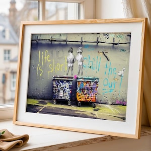 Può includere: Stampa artistica incorniciata con un muro coperto di graffiti con le parole "life is short" e "Chill the Out". Due bidoni colorati sono di fronte al muro. L'opera d'arte è esposta su un davanzale.