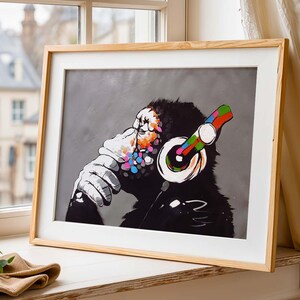May include: Framed art print of a monkey wearing headphones. The artwork uses a black and white palette with colourful accents on the face and headphones. The print is set against a grey background and framed in natural wood.
