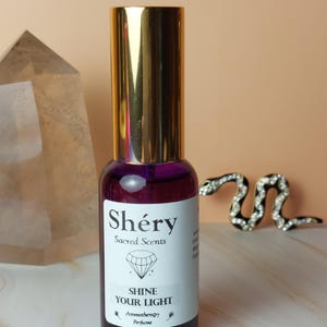 Può includere: Un flacone di profumo in vetro viola con tappo dorato. L'etichetta recita "Shéry Sacred Scents" e "Shine Your Light". Il flacone è per aromaterapia e profumo. Un cristallo e un serpente decorativo sono sullo sfondo.
