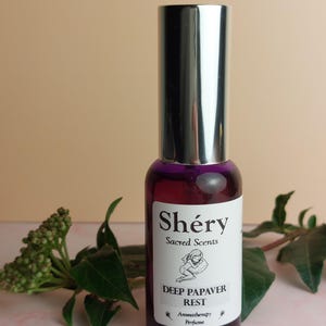 Könnte beinhalten: Eine lila Glas-Parfümflasche mit silberner Kappe. Das Etikett lautet "Shéry Sacred Scents" und "Deep Papaver Rest" mit einer Illustration einer ruhenden Person. Grüne Blätter umgeben die Flasche.