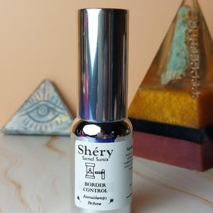 Può includere: Un flacone di profumo con tappo argentato con la scritta "Shéry Sacred Scents Border Control Aromatherapy Perfume". Il flacone è su una superficie chiara, con forme di piramide decorative sullo sfondo.