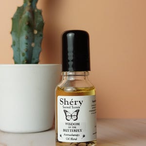 Op de afbeelding: Aromatherapie-olieblend in een heldere glazen fles met een zwarte dop. Het etiket luidt "Shéry Sacred Scents" en "Wisdom of the Butterfly". Een cactus in een witte pot staat op de achtergrond.
