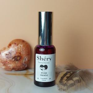 Op de afbeelding: Een kleine, donkerrode parfumfles met een zilveren dop. Het etiket luidt "Shéry Sacred Scents" en "POTJE TROOST". Een hartvormige steen en een veer staan op de achtergrond.