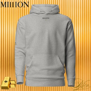 Puede incluir: Una sudadera con capucha gris con la palabra "MILLION" impresa en negro en el pecho y la manga izquierda. La sudadera tiene un bolsillo delantero y una capucha con cordón. El fondo es un patrón de rayas verticales de color dorado.