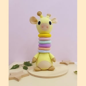May include: A yellow plush giraffe toy with a stack of colorful rings for a neck. The giraffe has brown ears, eyes, and a pink nose. The toy is sitting on a wooden base with star-shaped decorations.