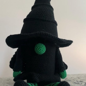 Pode incluir: Uma boneca de gnomo de crochê feita à mão com uma fantasia de bruxa. O gnomo é principalmente preto com um chapéu pontudo, um nariz verde e detalhes verdes nas mãos e nos pés. A boneca está sentada em uma superfície estampada.