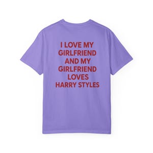 Puede incluir: Una camiseta de manga corta de color morado claro con texto rojo impreso en la parte posterior que dice: I LOVE MY GIRLFRIEND AND MY GIRLFRIEND LOVES HARRY STYLES. La prenda tiene cuello redondo y un corte est&aacute;ndar, mostrando la parte trasera.