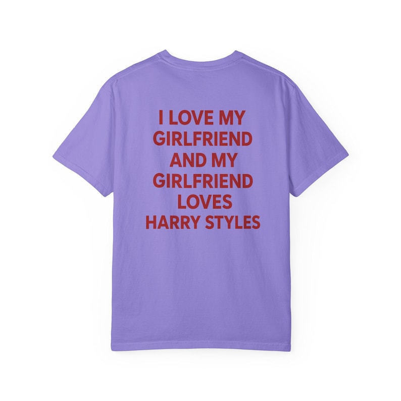 Puede incluir: Una camiseta de manga corta de color morado claro con texto rojo impreso en la parte posterior que dice: I LOVE MY GIRLFRIEND AND MY GIRLFRIEND LOVES HARRY STYLES. La prenda tiene cuello redondo y un corte est&aacute;ndar, mostrando la parte trasera.