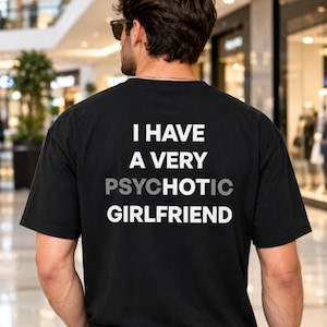 Op de afbeelding: Een zwart t-shirt met korte mouwen en witte tekst op de achterkant die luidt I HAVE A VERY PSYCHOTIC GIRLFRIEND, waarbij de letters HOT in een lichtere grijze kleur in het woord PSYCHOTIC zijn gemarkeerd om het woord HOT te benadrukken.