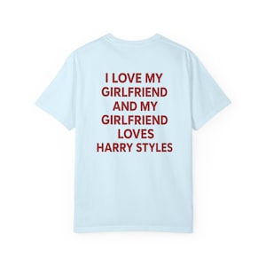 Puede incluir: Una camiseta de manga corta de color azul claro con texto rojo oscuro impreso en la parte posterior que dice: I LOVE MY GIRLFRIEND AND MY GIRLFRIEND LOVES HARRY STYLES. La prenda tiene cuello redondo y corte est&aacute;ndar, mostrada solo de espaldas.