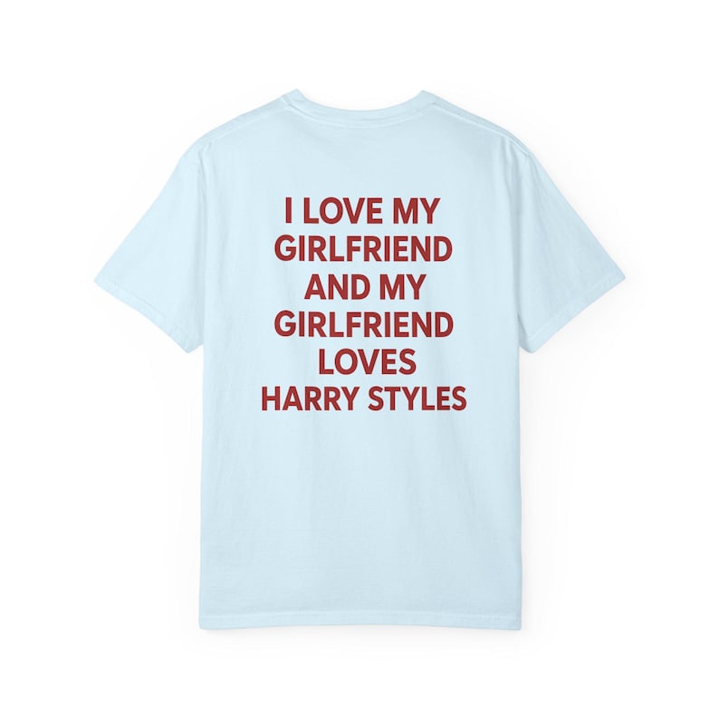Puede incluir: Una camiseta de manga corta de color azul claro con texto rojo oscuro impreso en la parte posterior que dice: I LOVE MY GIRLFRIEND AND MY GIRLFRIEND LOVES HARRY STYLES. La prenda tiene cuello redondo y corte est&aacute;ndar, mostrada solo de espaldas.