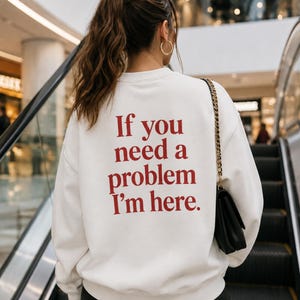 Pode incluir: Uma camisola branca de manga comprida com gola redonda e texto vermelho nas costas que diz: "If you need a problem I'm here." A peça tem gola, punhos e bainha canelados, e é usada com uma mala de ombro preta com corrente dourada.