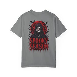 以下が含まれることがあります： グレーの半袖Tシャツ。背中には白いマスクと赤い目をしたフード付きの人物のグラフィックが描かれています。背景には大きな赤い月と黒いコウモリが配置されています。赤い文字で「SPOOKY SEASON」とプリントされており、滴るようなエフェクトが施されています。