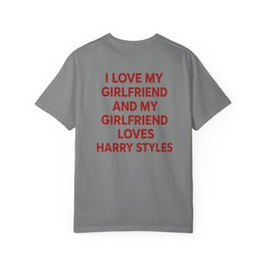 Puede incluir: Una camiseta gris de manga corta con texto rojo impreso en la parte posterior que dice: I LOVE MY GIRLFRIEND AND MY GIRLFRIEND LOVES HARRY STYLES. La prenda tiene cuello redondo y un corte est&aacute;ndar, mostrando la vista trasera de la camiseta.