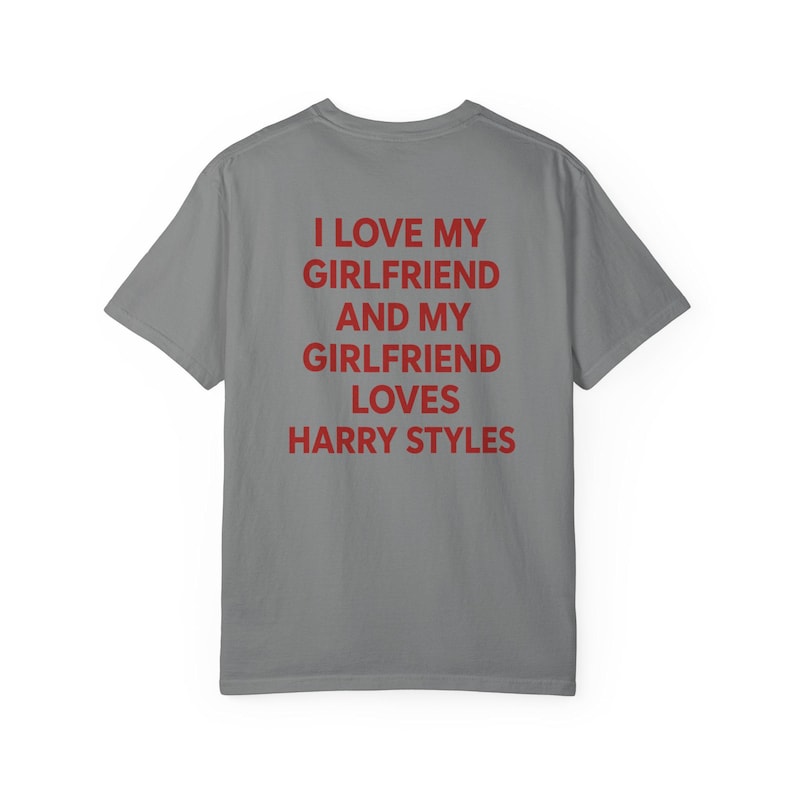 Puede incluir: Una camiseta gris de manga corta con texto rojo impreso en la parte posterior que dice: I LOVE MY GIRLFRIEND AND MY GIRLFRIEND LOVES HARRY STYLES. La prenda tiene cuello redondo y un corte est&aacute;ndar, mostrando la vista trasera de la camiseta.