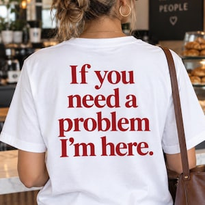 May include: A white short-sleeved t-shirt featuring red text printed on the back that reads, "If you need a problem I'm here." The shirt has a crew neckline and a standard fit, shown from the back view with a brown shoulder bag strap visible.
