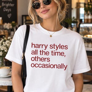 May include: A white short-sleeved t-shirt featuring the red text "harry styles all the time, others occasionally" printed on the front. The shirt has a crew neckline and a relaxed fit, worn with a black shoulder bag and gold-toned jewelry.