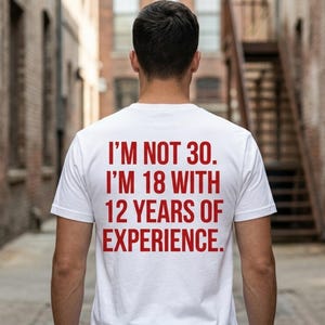 May include: A white short-sleeved t-shirt featuring red text on the back that reads: I'M NOT 30. I'M 18 WITH 12 YEARS OF EXPERIENCE. The shirt has a crew neckline and is made of a plain white fabric with bold, capitalised red lettering.