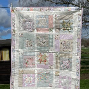 May include: A patchwork quilt in pastel shades, including pink, green, and cream. The quilt is made up of various fabric patterns, including floral and polka dot designs. The quilt is hanging on a washing line, with a light pink border.