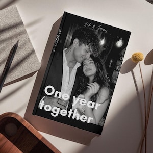 Può includere: Libro fotografico in bianco e nero con la scritta "One year together" e i nomi "Ash & Jenny". La copertina presenta una coppia che si abbraccia. Sono visibili anche una matita, un taccuino e fiori secchi.